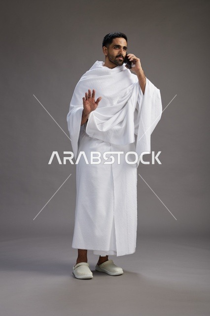 A portrait of a Saudi man in Ihram clothing making a phone call on a mobile phone, preparing to perform the rituals of Hajj and Umrah, full-body shot, gray background.