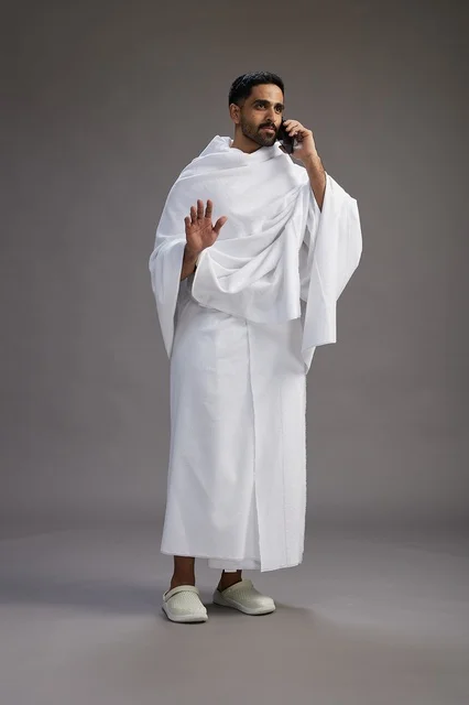 A portrait of a Saudi man in Ihram clothing making a phone call on a mobile phone, preparing to perform the rituals of Hajj and Umrah, full-body shot, gray background. A portrait of a Saudi man in Ihram clothing making a phone call on a mobile phone, preparing to perform the rituals of Hajj and Umrah, full-body shot, gray background.