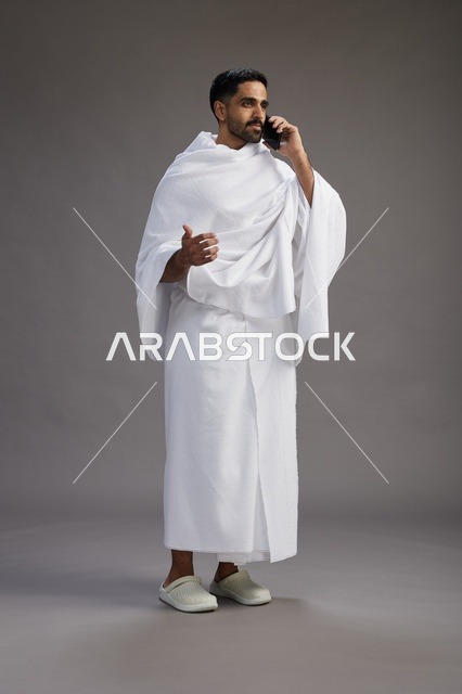بورتريه لرجل سعودي بملابس الإحرام يجري مكالمة هاتفية عبر الهاتف المحمول, الاستعداد لاداء مناسك الحج والعمره, الجسم بالطول الكامل , خلفيه رماديه