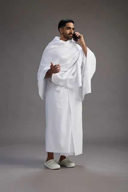 A portrait of a Saudi man in Ihram clothing making a phone call on a mobile phone, preparing to perform the rituals of Hajj and Umrah, full-body shot, gray background.