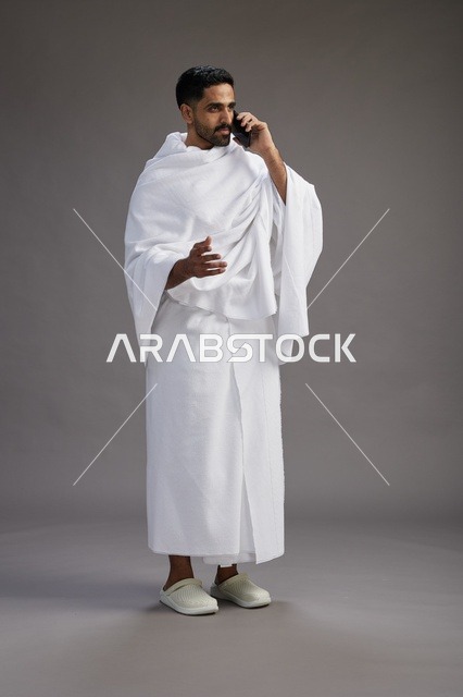 بورتريه لرجل سعودي بملابس الإحرام يجري مكالمة هاتفية عبر الهاتف المحمول, الاستعداد لاداء مناسك الحج والعمره, الجسم بالطول الكامل , خلفيه رماديه