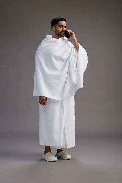 A portrait of a Saudi man in Ihram clothing making a phone call on a mobile phone, preparing to perform the rituals of Hajj and Umrah, full-body shot, gray background.