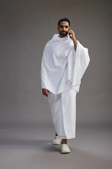 A portrait of a Saudi man in Ihram clothing making a phone call on a mobile phone, preparing to perform the rituals of Hajj and Umrah, full-body shot, gray background.