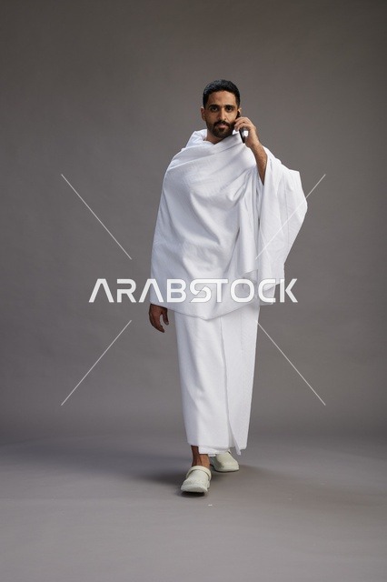 بورتريه لرجل سعودي بملابس الإحرام يجري مكالمة هاتفية عبر الهاتف المحمول, الاستعداد لاداء مناسك الحج والعمره, الجسم بالطول الكامل , خلفيه رماديه