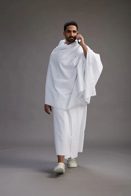 A portrait of a Saudi man in Ihram clothing making a phone call on a mobile phone, preparing to perform the rituals of Hajj and Umrah, full-body shot, gray background. A portrait of a Saudi man in Ihram clothing making a phone call on a mobile phone, preparing to perform the rituals of Hajj and Umrah, full-body shot, gray background.