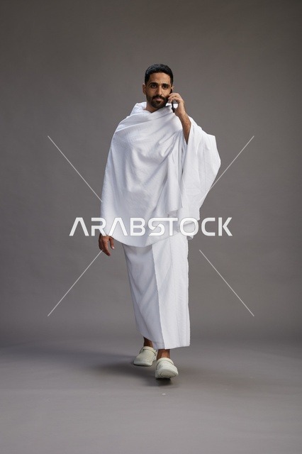 بورتريه لرجل سعودي بملابس الإحرام يجري مكالمة هاتفية عبر الهاتف المحمول, الاستعداد لاداء مناسك الحج والعمره, الجسم بالطول الكامل , خلفيه رماديه