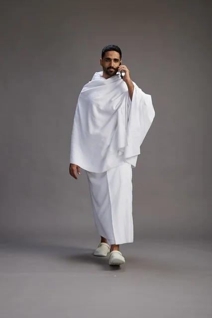 A portrait of a Saudi man in Ihram clothing making a phone call on a mobile phone, preparing to perform the rituals of Hajj and Umrah, full-body shot, gray background.