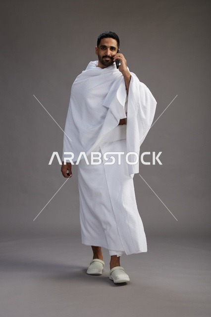 بورتريه لرجل سعودي بملابس الإحرام يجري مكالمة هاتفية عبر الهاتف المحمول, الاستعداد لاداء مناسك الحج والعمره, الجسم بالطول الكامل , خلفيه رماديه