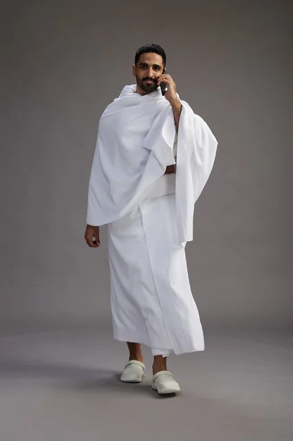A portrait of a Saudi man in Ihram clothing making a phone call on a mobile phone, preparing to perform the rituals of Hajj and Umrah, full-body shot, gray background.