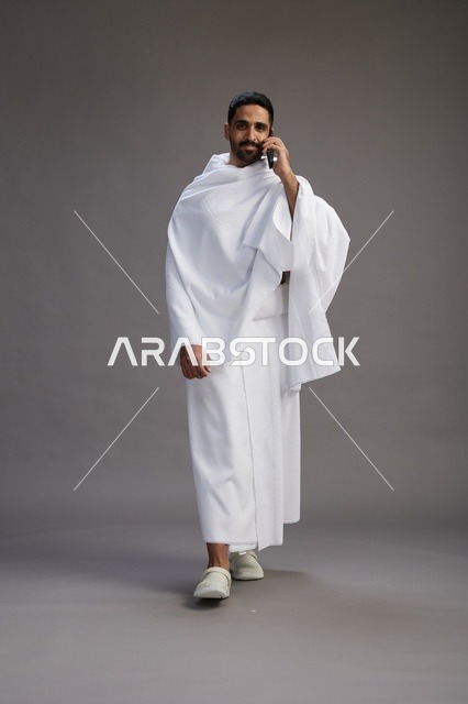 بورتريه لرجل سعودي بملابس الإحرام يجري مكالمة هاتفية عبر الهاتف المحمول, الاستعداد لاداء مناسك الحج والعمره, الجسم بالطول الكامل , خلفيه رماديه