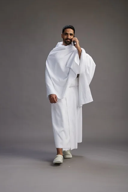 A portrait of a Saudi man in Ihram clothing making a phone call on a mobile phone, preparing to perform the rituals of Hajj and Umrah, full-body shot, gray background. A portrait of a Saudi man in Ihram clothing making a phone call on a mobile phone, preparing to perform the rituals of Hajj and Umrah, full-body shot, gray background.