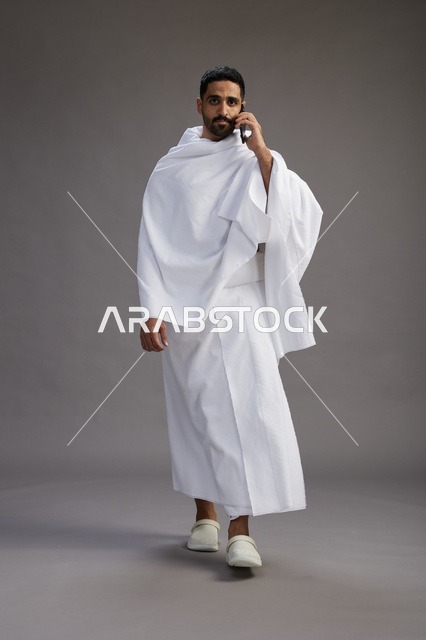 A portrait of a Saudi man in Ihram clothing making a phone call on a mobile phone, preparing to perform the rituals of Hajj and Umrah, full-body shot, gray background.