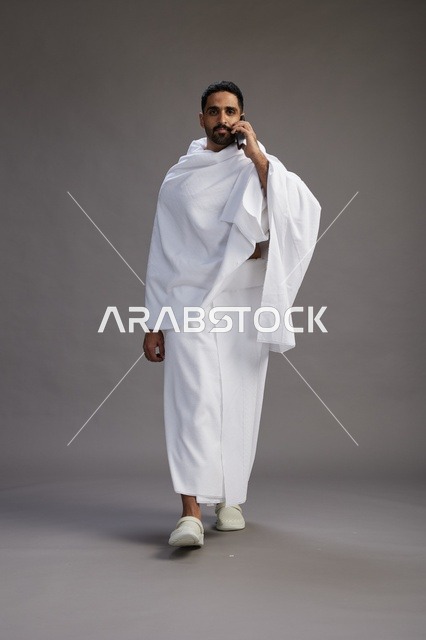 A portrait of a Saudi man in Ihram clothing making a phone call on a mobile phone, preparing to perform the rituals of Hajj and Umrah, full-body shot, gray background.