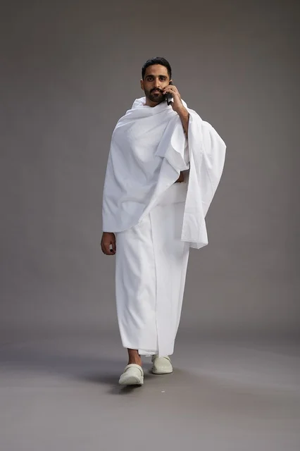بورتريه لرجل سعودي بملابس الإحرام يجري مكالمة هاتفية عبر الهاتف المحمول, الاستعداد لاداء مناسك الحج والعمره, الجسم بالطول الكامل , خلفيه رماديه