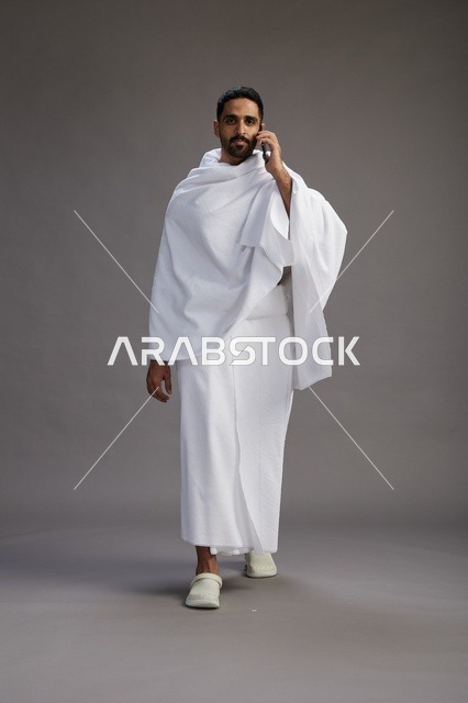 بورتريه لرجل سعودي بملابس الإحرام يجري مكالمة هاتفية عبر الهاتف المحمول, الاستعداد لاداء مناسك الحج والعمره, الجسم بالطول الكامل , خلفيه رماديه
