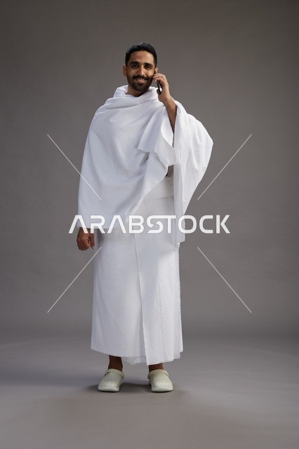 بورتريه لرجل سعودي بملابس الإحرام يجري مكالمة هاتفية عبر الهاتف المحمول, الاستعداد لاداء مناسك الحج والعمره, الجسم بالطول الكامل , خلفيه رماديه