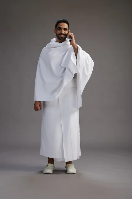 A portrait of a Saudi man in Ihram clothing making a phone call on a mobile phone, preparing to perform the rituals of Hajj and Umrah, full-body shot, gray background.