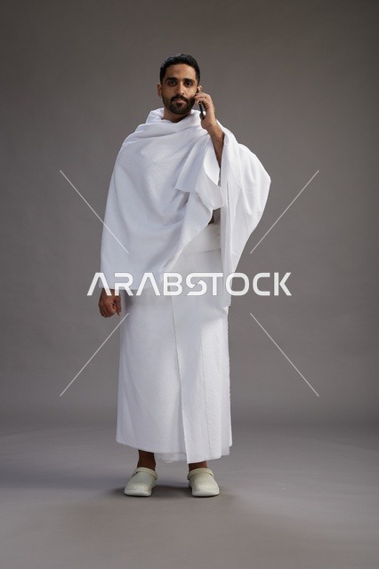 بورتريه لرجل سعودي بملابس الإحرام يجري مكالمة هاتفية عبر الهاتف المحمول, الاستعداد لاداء مناسك الحج والعمره, الجسم بالطول الكامل , خلفيه رماديه
