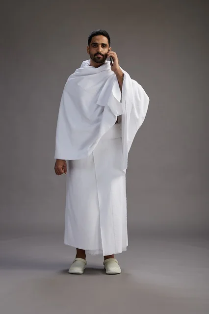 A portrait of a Saudi man in Ihram clothing making a phone call on a mobile phone, preparing to perform the rituals of Hajj and Umrah, full-body shot, gray background. A portrait of a Saudi man in Ihram clothing making a phone call on a mobile phone, preparing to perform the rituals of Hajj and Umrah, full-body shot, gray background.