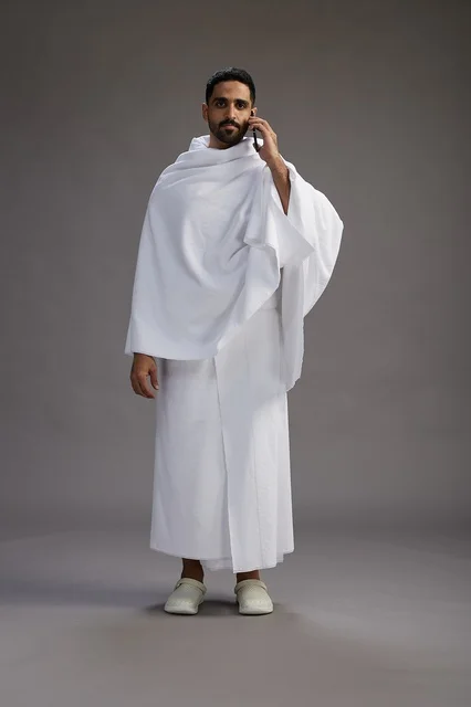 A portrait of a Saudi man in Ihram clothing making a phone call on a mobile phone, preparing to perform the rituals of Hajj and Umrah, full-body shot, gray background. A portrait of a Saudi man in Ihram clothing making a phone call on a mobile phone, preparing to perform the rituals of Hajj and Umrah, full-body shot, gray background.