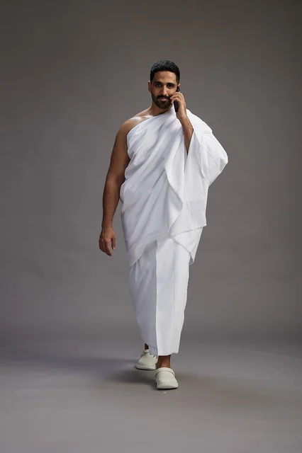 A portrait of a Saudi man in Ihram clothing making a phone call on a mobile phone, preparing to perform the rituals of Hajj and Umrah, full-body shot, gray background.