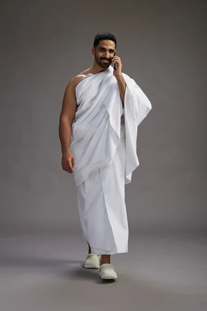 A portrait of a Saudi man in Ihram clothing making a phone call on a mobile phone, preparing to perform the rituals of Hajj and Umrah, full-body shot, gray background. A portrait of a Saudi man in Ihram clothing making a phone call on a mobile phone, preparing to perform the rituals of Hajj and Umrah, full-body shot, gray background.