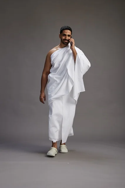 A portrait of a Saudi man in Ihram clothing making a phone call on a mobile phone, preparing to perform the rituals of Hajj and Umrah, full-body shot, gray background. A portrait of a Saudi man in Ihram clothing making a phone call on a mobile phone, preparing to perform the rituals of Hajj and Umrah, full-body shot, gray background.
