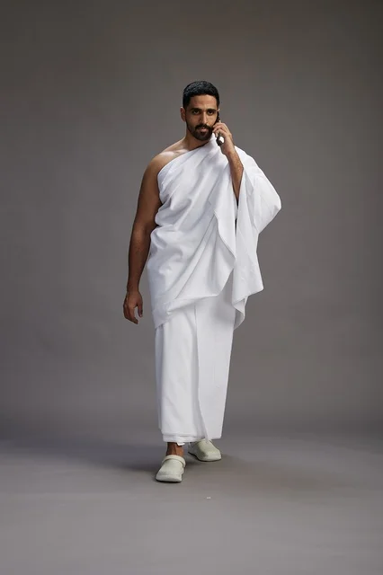 A portrait of a Saudi man in Ihram clothing making a phone call on a mobile phone, preparing to perform the rituals of Hajj and Umrah, full-body shot, gray background.
