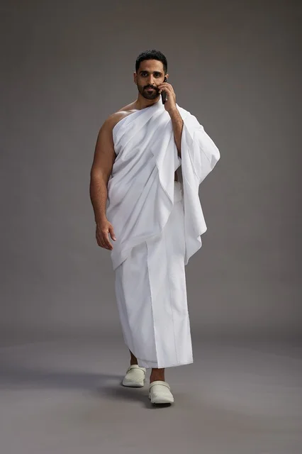 A portrait of a Saudi man in Ihram clothing making a phone call on a mobile phone, preparing to perform the rituals of Hajj and Umrah, full-body shot, gray background.