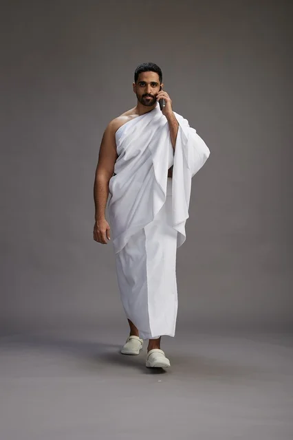 A portrait of a Saudi man in Ihram clothing making a phone call on a mobile phone, preparing to perform the rituals of Hajj and Umrah, full-body shot, gray background.