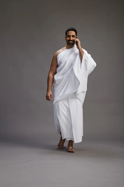 A portrait of a Saudi man in Ihram clothing making a phone call on a mobile phone, preparing to perform the rituals of Hajj and Umrah, full-body shot, gray background.