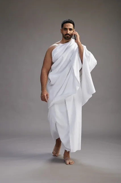 A portrait of a Saudi man in Ihram clothing making a phone call on a mobile phone, preparing to perform the rituals of Hajj and Umrah, full-body shot, gray background. A portrait of a Saudi man in Ihram clothing making a phone call on a mobile phone, preparing to perform the rituals of Hajj and Umrah, full-body shot, gray background.