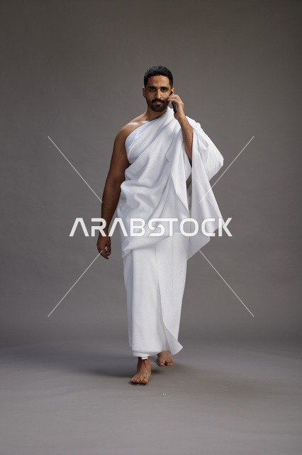A portrait of a Saudi man in Ihram clothing making a phone call on a mobile phone, preparing to perform the rituals of Hajj and Umrah, full-body shot, gray background.