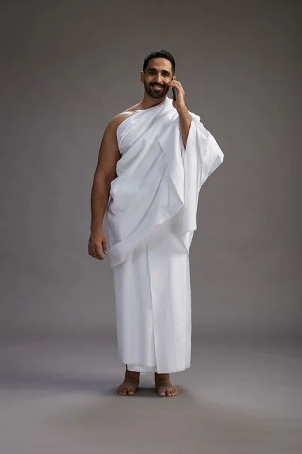 بورتريه لرجل سعودي بملابس الإحرام يجري مكالمة هاتفية عبر الهاتف المحمول, الاستعداد لاداء مناسك الحج والعمره, الجسم بالطول الكامل , خلفيه رماديه