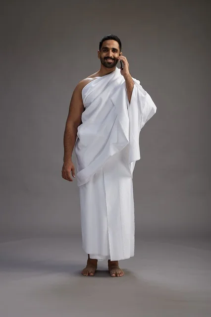 A portrait of a Saudi man in Ihram clothing making a phone call on a mobile phone, preparing to perform the rituals of Hajj and Umrah, full-body shot, gray background.