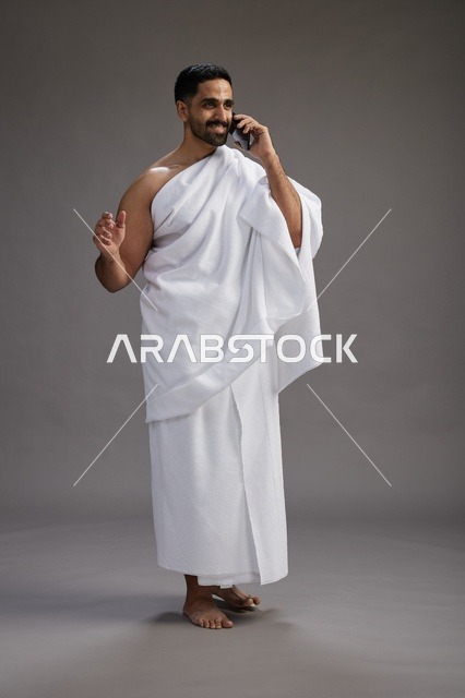 A portrait of a Saudi man in Ihram clothing making a phone call on a mobile phone, preparing to perform the rituals of Hajj and Umrah, full-body shot, gray background.