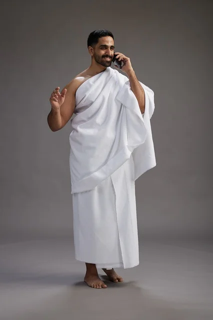A portrait of a Saudi man in Ihram clothing making a phone call on a mobile phone, preparing to perform the rituals of Hajj and Umrah, full-body shot, gray background.