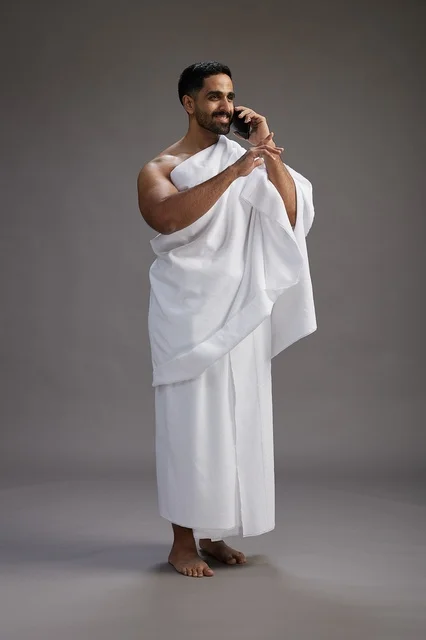 بورتريه لرجل سعودي بملابس الإحرام يجري مكالمة هاتفية عبر الهاتف المحمول, الاستعداد لاداء مناسك الحج والعمره, الجسم بالطول الكامل , خلفيه رماديه