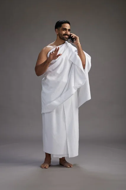 A portrait of a Saudi man in Ihram clothing making a phone call on a mobile phone, preparing to perform the rituals of Hajj and Umrah, full-body shot, gray background.