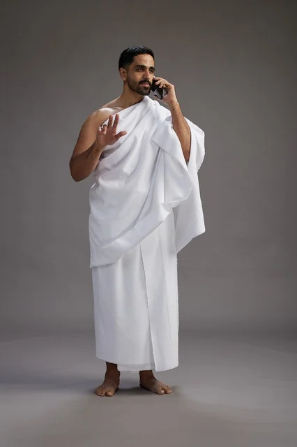 A portrait of a Saudi man in Ihram clothing making a phone call on a mobile phone, preparing to perform the rituals of Hajj and Umrah, full-body shot, gray background. A portrait of a Saudi man in Ihram clothing making a phone call on a mobile phone, preparing to perform the rituals of Hajj and Umrah, full-body shot, gray background.