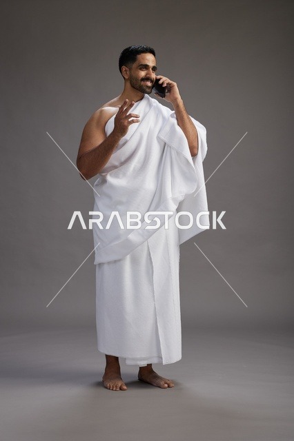 A portrait of a Saudi man in Ihram clothing making a phone call on a mobile phone, preparing to perform the rituals of Hajj and Umrah, full-body shot, gray background.