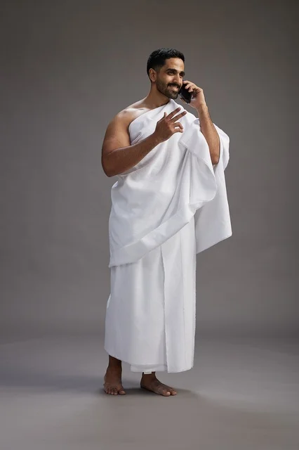 A portrait of a Saudi man in Ihram clothing making a phone call on a mobile phone, preparing to perform the rituals of Hajj and Umrah, full-body shot, gray background. A portrait of a Saudi man in Ihram clothing making a phone call on a mobile phone, preparing to perform the rituals of Hajj and Umrah, full-body shot, gray background.