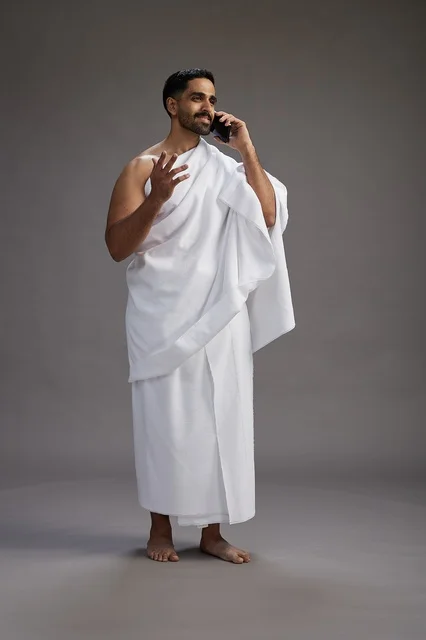 بورتريه لرجل سعودي بملابس الإحرام يجري مكالمة هاتفية عبر الهاتف المحمول, الاستعداد لاداء مناسك الحج والعمره, الجسم بالطول الكامل , خلفيه رماديه