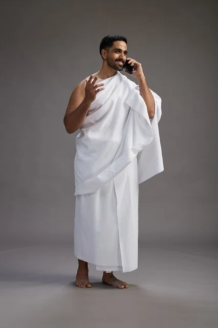 بورتريه لرجل سعودي بملابس الإحرام يجري مكالمة هاتفية عبر الهاتف المحمول, الاستعداد لاداء مناسك الحج والعمره, الجسم بالطول الكامل , خلفيه رماديه