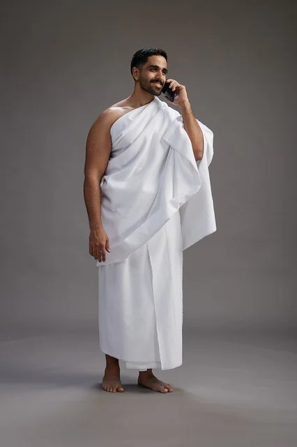 A portrait of a Saudi man in Ihram clothing making a phone call on a mobile phone, preparing to perform the rituals of Hajj and Umrah, full-body shot, gray background.