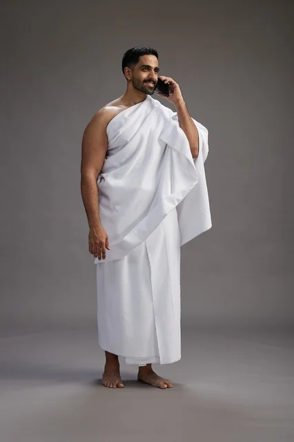 A portrait of a Saudi man in Ihram clothing making a phone call on a mobile phone, preparing to perform the rituals of Hajj and Umrah, full-body shot, gray background.
