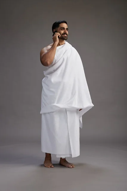 A portrait of a Saudi man in Ihram clothing making a phone call on a mobile phone, preparing to perform the rituals of Hajj and Umrah, full-body shot, gray background.