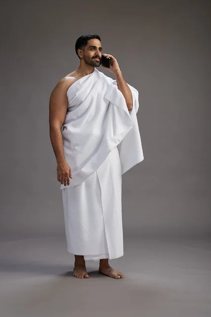 بورتريه لرجل سعودي بملابس الإحرام يجري مكالمة هاتفية عبر الهاتف المحمول, الاستعداد لاداء مناسك الحج والعمره, الجسم بالطول الكامل , خلفيه رماديه