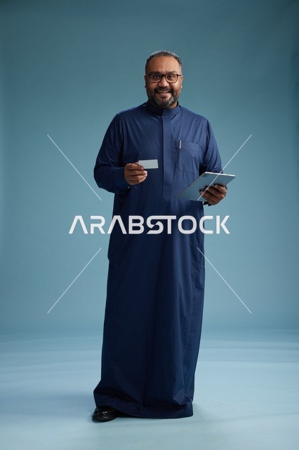 بورتريه لرجل سعودي يرتدي ثوباً أزرق يمسك بيده بطاقة بيضاء فارغه مستخدماً جهازاً لوحياً, الدفع الالكتروني عبر البطاقة البنكيه, الجسم بالطول الكامل, خلفيه زرقاء