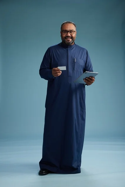 A portrait of a Saudi man wearing a blue thobe holding a blank white card in one hand while using a tablet, electronic payment via a bank card, full body, blue background.